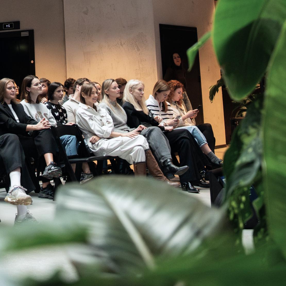 Nord Security employees seated in an audience at a company talk, seen past a leafy plant.