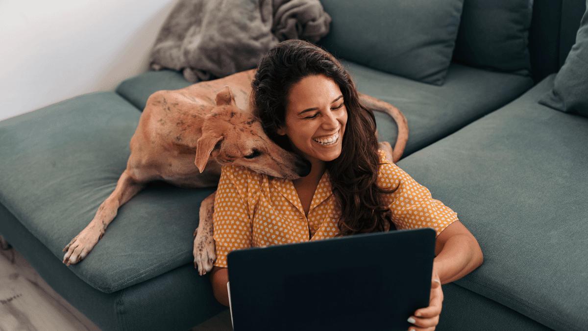 Woman using a laptop on the sofa with her dog beside her.
