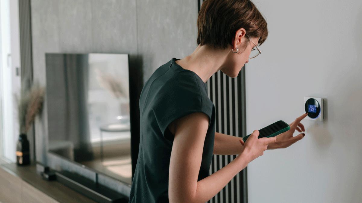 A woman holding a smartphone adjusting the temperature of her room on a termostat.