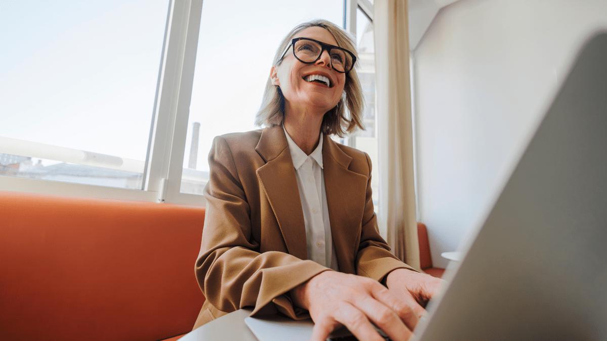 Woman smiling while working at a laptop by a window.