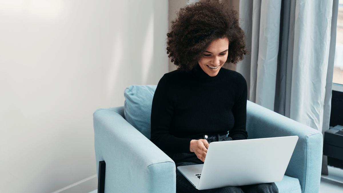 Woman smiling at her laptop while sitting in an armchair.